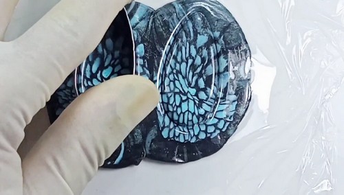 A gloved hand pressing a clear oval cutter into the black and teal patterned polymer clay sheet on a plastic-wrapped surface to cut earring shapes.