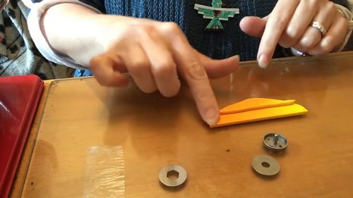 Hands folding a flat strip of orange polymer clay on a wooden board beside two metal washer cutters.