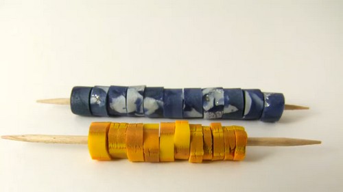 Two toothpick skewers holding rows of blue marbled and yellow polymer clay heishi beads before baking.