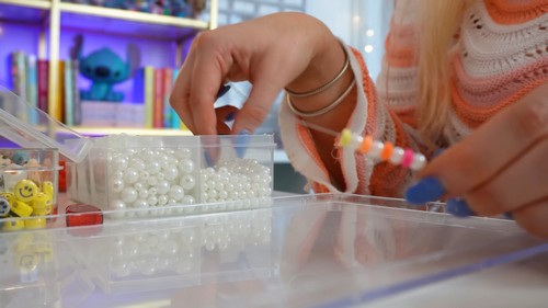 A hand reaching into a container of white pearl beads while holding a partly finished clay bead bracelet