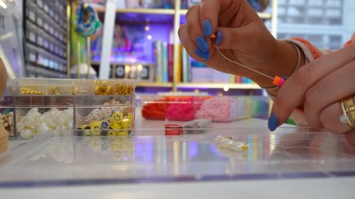 Hands threading a strand of colorful clay beads onto stretch cord at a bead studio table