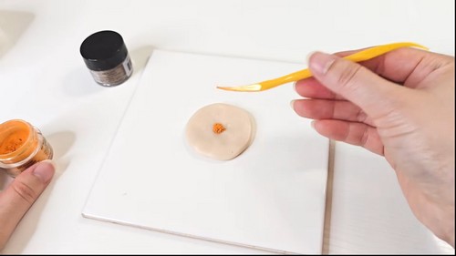 Brush applying a small dot of orange powder pigment to a round flattened clay disc on a white tile