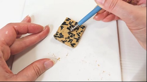 Clay tool pressing black fragment pieces into a square of gold base clay to begin building the tortoise shell pattern