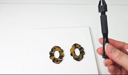 Two flower shaped cutouts of tortoise shell patterned clay with open centers resting on a white tile beside a hole punch tool