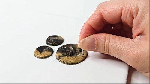Hand piercing a small hole in one of three black and gold circular clay pieces with a needle tool