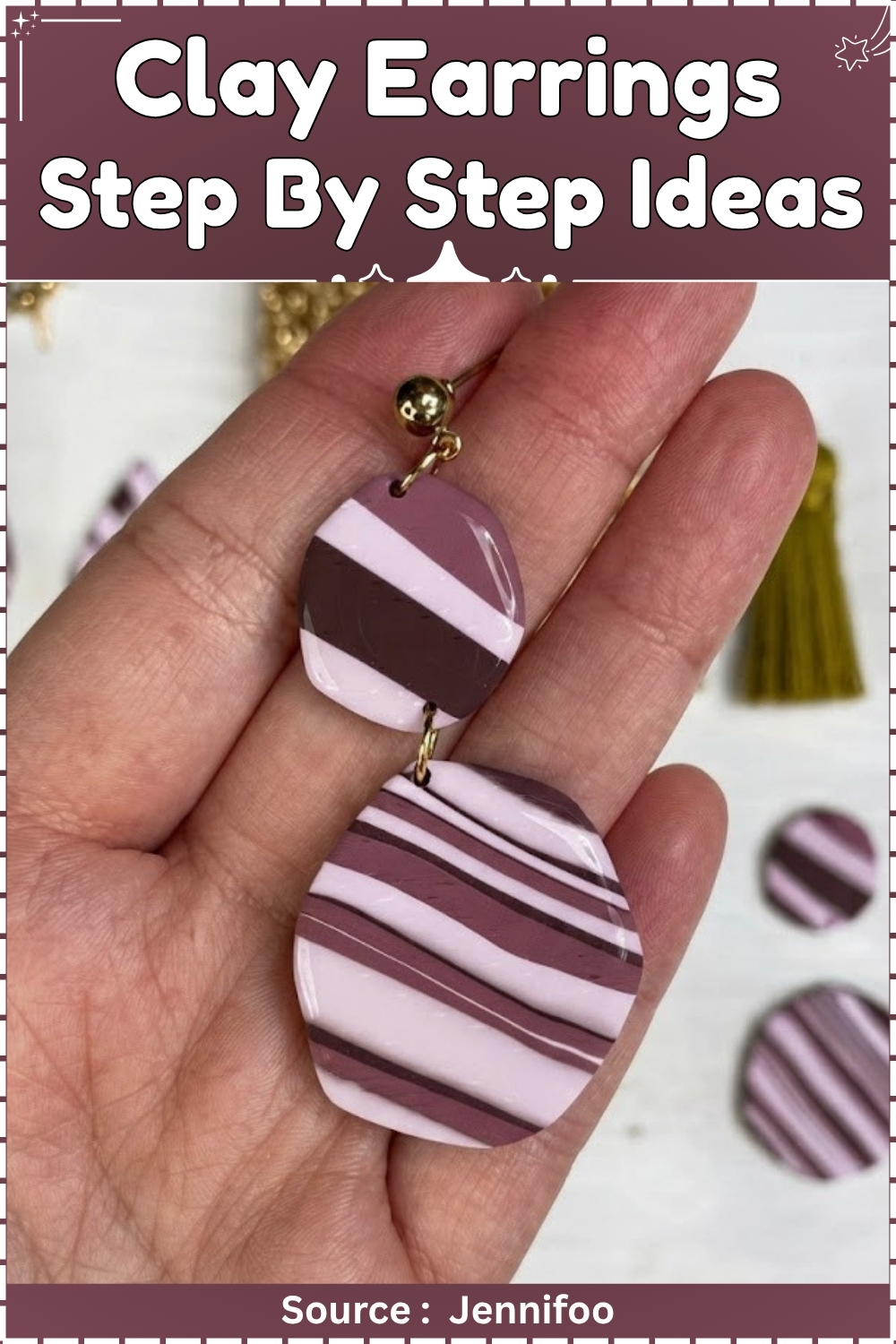 A hand holding a finished pair of two-tier polymer clay earrings in pink, mauve, and dark burgundy diagonal stripes connected with a gold jump ring and gold ball stud. 