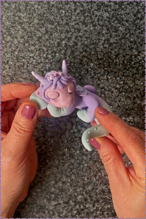 Hands holding a finished lavender and mint polymer clay unicorn figurine with a twisted horn, pearl bead hair details, and a mint crescent tail, shown against a gray gravel surface.