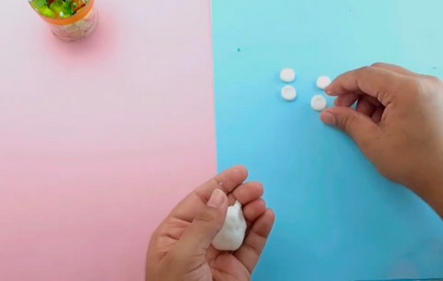 Hands rolling a small piece of white clay into a ball with three flat white discs already formed on a pink and blue surface