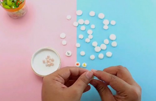 Hands threading a needle through small clay letter beads near a small dish of silver beads with blank white discs scattered behind